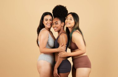 Değişik milletlerden üç kadın sarılıyor, doğal güzellikleri yayıyor ve bej arka planda rahat iç çamaşırlarıyla kadınlığın çeşitliliğini ve gücünü sergiliyor.