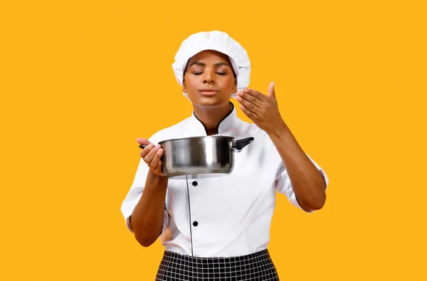 Delicious Smell. Black Chef Woman In Uniform Holding Saucepan And Smelling Meal, Professional Cook Lady Standing Isolated Over Yellow Studio Background, Enjoying Cooking, Copy Space