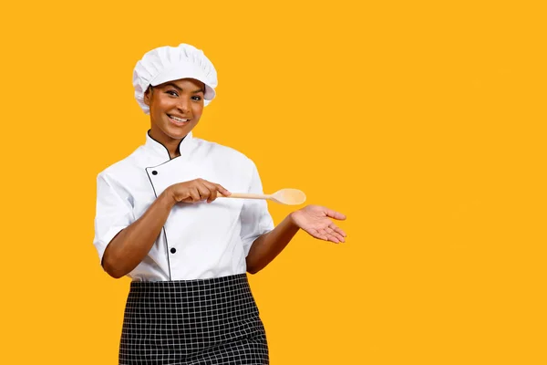 Smiling Black Chef Woman With Wooden Spatula Pointing At Her Empty Palm, Happy African American Cook Lady In Uniform Presenting Invisible Object, Standing Isolated On Yellow Background, Copy Space