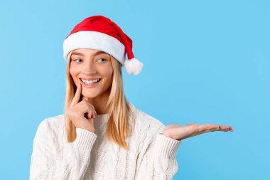 Noel Baba şapkalı mutlu genç kadın parmaklarını dudaklarına dayayıp gülümsüyor, sanki cazip bir kış Noel indirimi ve mevsimsel teklifler sunuyormuş gibi elini açık tutuyor.