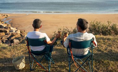 Coastal Coffee Anları. Üst düzey eşler ellerinde kahve fincanlarıyla sandalyelerde dinleniyor, okyanus dalgalarını izliyor, yaz günü deniz kenarında hafta sonlarını dışarıda geçiriyorlar. Arkadan görüntüle