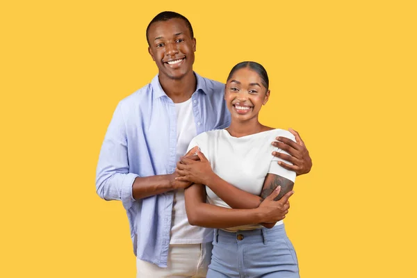 Loving black man hugging his wife from back, standing and smiling together over yellow studio background. Happy guy and lady posing to camera
