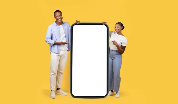 stock image Cool mobile offer. Happy black man and woman pointing at huge cellphone with white screen, demonstrating free space for phone app or website design, yellow background