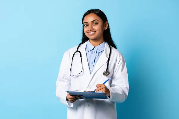 Professional Indian lady physician with stethoscope and medical chart, posing in studio on blue background, smiling to camera. Healthcare career and expertise concept