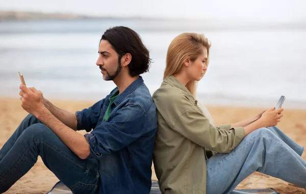 Young man and woman sitting back to back and using smartphones outdoors, millennial couple checking social media or browsing internet during date on the beach, suffering gadget addiction