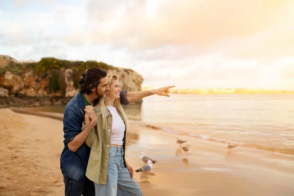 Romantic Man And Woman Relaxing On The Beach At Sunset Time, Beautiful Young Couple Standing Outdoors At Sea Shore, Embracing And Smiling, Enjoying Date Outside, Man Pointing Away With Hand