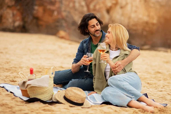 Portrait Of Romantic Young Couple Having Date Outdoors, Drinking Wine And Embracing, Loving Millennial Man And Woman Enjoying Picnic On The Beach, Clinking Glasses And Smiling To Each Other