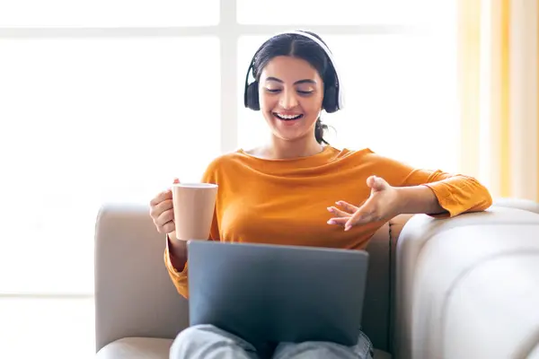 Happy young indian woman making video call via laptop and drinking coffee at home, smiling eastern lady wearing wireless headphones and gesturing at webcamera, sitting on sofa in living room
