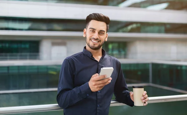 Arab entrepreneur man with smartphone and coffee paper cup checking messages on a business app, posing near urban office building, smiling to camera. Gadgets lifestyle