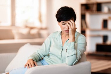 Stres ve baş ağrısı. Orta yaşlı bir kadın oturma odasındaki koltukta otururken alnına sürtünüyor, şiddetli migren ağrısı çekiyor evde otururken başı ağrıyor. Sağlık sorunları kavramı