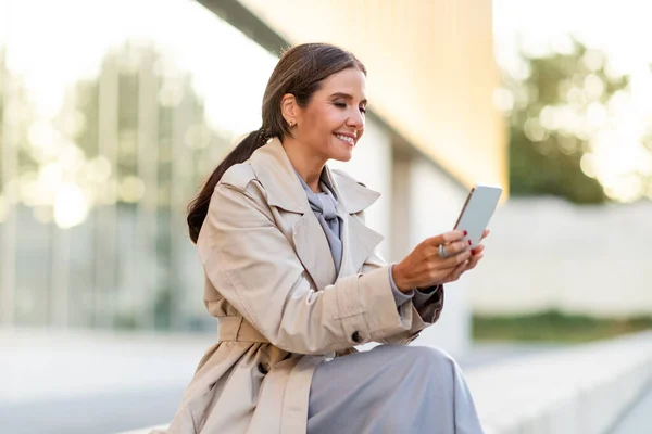 Mature lady manager have break, sitting on bench by office building, smiling at smartphone screen, enjoying video call with family, copy space. Businesswoman using cell phone outdoors