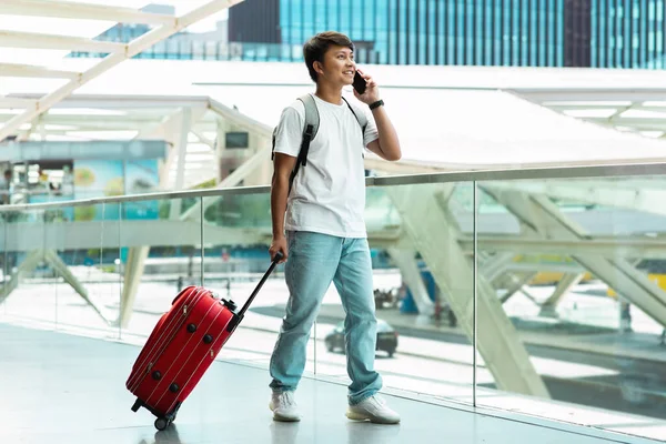 Cheerful young asian man tourist walking by sirport, carrying luggage, talking on phone with friend, calling taxi, arrived to new city, chinese guy traveling, copy space, full length