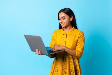 Mutlu genç Hindu bayan dizüstü bilgisayarını geniş web sörfü, online araştırma ve modern e-öğrenme çevrimiçi oturumlarında mavi arka planda poz vermek için kullanıyor. Stüdyo çekimi