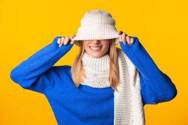 Neşeli kadın beyaz örgü şapkasını indirir ve gülümser, mavi süveter ve beyaz eşarp giyer, parlak sarı arka plana yaslanır.