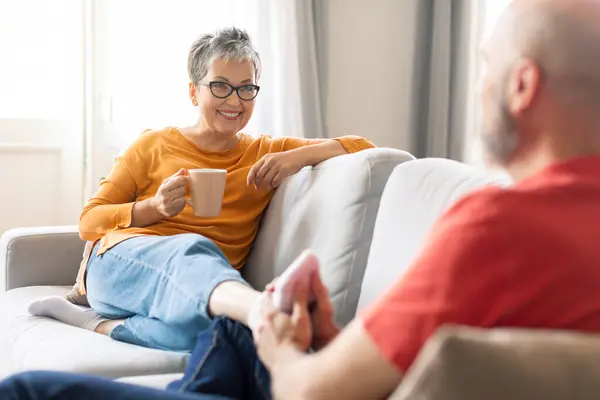 Loving senior husband making foot massage for his wife at home, helping her to relax, happy elderly spouses resting on couch in living room together, woman drinking coffee, selective focus