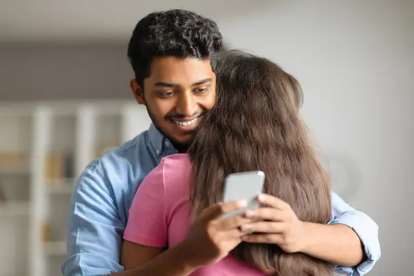 Smiling young Indian man embraces his wife, discreetly using his cellphone, hinting at possible infidelity, relaxed home setting background