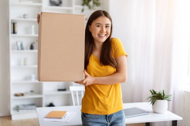 Teslimat Konsepti. Mutlu Asyalı kadın, elinde kocaman bir karton kutuyla kameraya gülümsüyor, neşeli, genç Çinli kadın, evinde oturma odasında dururken paketi teslim ediyor.