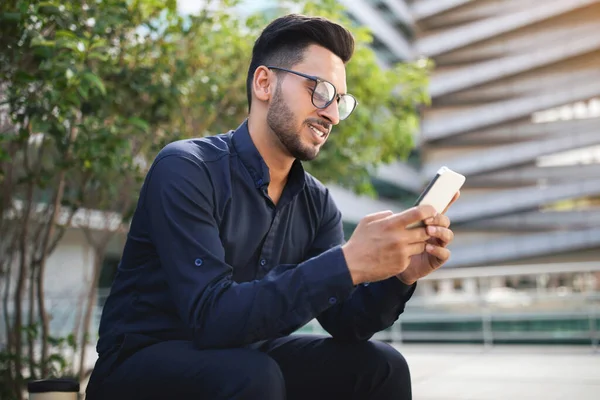 Arab entrepreneur man in glasses holding cellphone and browsing new online platform, sitting near modern office building in urban setting. Corporate communication and tech advancements concept