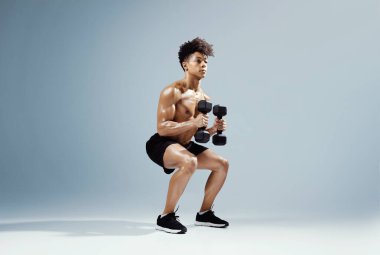 Kararlı sporcu, dambılları sıkıca kavrıyor, ağırlığını gri stüdyo arka planına vererek egzersiz yapıyor. Güç antrenmanı ve disiplin. Tam boy üstsüz kaslı fitness adamı.