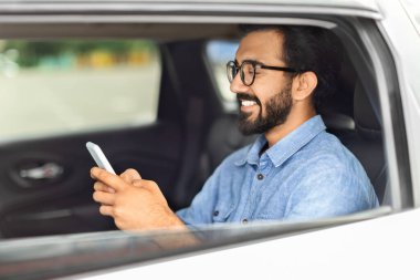 Genç Hintli işadamı eve arabayla giderken e-postaları kontrol ediyor, arka koltukta oturuyor ve cep telefonu kullanıyor, kopyalama alanı kullanıyor. Sıradan giyinen, taksiyle gezen, akıllı telefon kullanan mutlu bir doğulu.