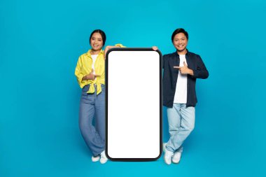 Mavi stüdyo arka planında izole edilmiş, boş yeri olan akıllı telefonları işaret eden mutlu Asyalı çift. Tavsiye aygıtı, sunum, reklam ve teklif