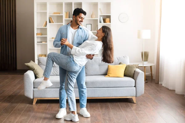 Young Indian couple, in casual attire, shares playful dance move in their homey living room, filled with natural light, contemporary furniture, full length, free space