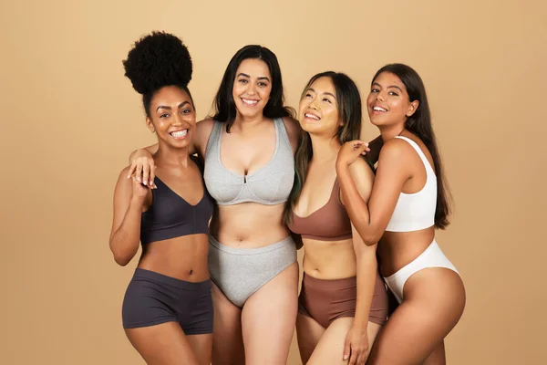 Four joyful, diverse women confidently pose in their underwear, celebrating natural beauty and camaraderie, embracing and smiling at camera on beige studio backdrop