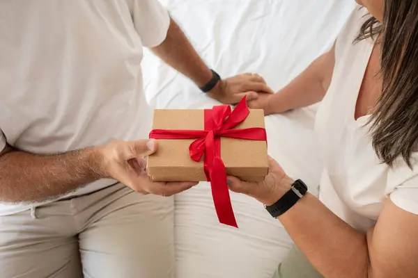 Positive senior caucasian woman gives gift box to man, sit on bad in bedroom interior, close up. Celebration birthday, anniversary surprise, holiday together, love and congratulate at home