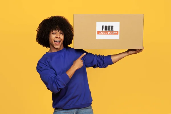 Happy Black Guy Pointing Finger At Cardboard Box With Free Delivery Text, Excited Young African American Man Recommending Shipping Services While Standing Isolated Over Yellow Studio Background