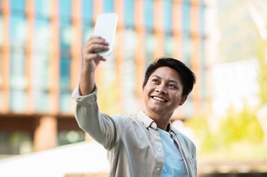 Sıradan kıyafetler içinde Asyalı genç ve mutlu girişimci sokakta akıllı telefondan selfie çekiyor. Çinli adam seyahat ederken fotoğraf çekiyor, sosyal medyada paylaşıyor, boşluğu kopyalıyor.