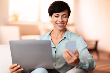 Evdeki modern oturma odasında otururken, dizüstü bilgisayar kullanırken cep telefonlu kulaklıklarla cep telefonuyla konuşan neşeli, orta yaşlı bir bayan. Modern iletişim ve serbest çalışma kavramı