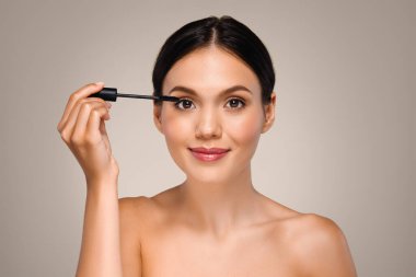 Mükemmel ciltli bin yıllık beyaz kadın kirpiklerine rimel sürüyor, çıplak temel makyajın keyfini çıkarıyor, gri stüdyo arka planında izole edilmiş. Güzellik hizmetleri, rutin prosedürler.