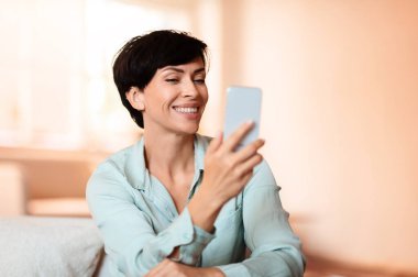 Orta yaşlı bir kadın internet üzerinden arama teklifleri sunuyor. Mobil uygulamalara dalmış, modern ev içi koltuklarda oturuyor. Dijital keşif kavramı ile boş zaman