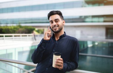 Orta Doğulu iş adamı cep telefonuyla konuşuyor modern şehir bölgesinde, ofis merkezinin yakınında siyah gömlekli, iletişim kurarken kameraya gülümsüyor. Meşgul şehir kurumsal yaşam tarzı