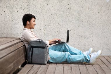 Milenyum Asyalı yakışıklı, uzaktan kumandalı, halka açık bir parkta çalışan, beton bankta oturan, dizüstü bilgisayarda, klavyede, yan görünümde, kopyalama alanında çalışan.