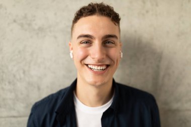 Sıradan, kablosuz kulaklıklı, güler yüzlü genç Avrupalı adam yaşam tarzını seviyor, gri duvar arka planında müzik dinliyor. Aygıt ve ses uygulamalı eğitim, kapatın