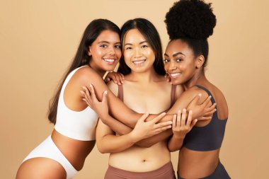 Farklı geçmişlere sahip üç genç kadın kameraya gülümsüyor, iç çamaşırlarıyla rahatça poz veriyor ve sarılıyorlar, bej arka planda doğal güzelliklerini sergiliyorlar..