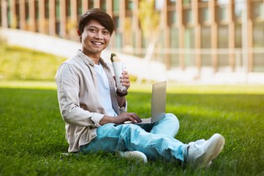 Mutlu yakışıklı Asyalı adam günlük kıyafetlerini giyiyor yeşil çimlerde oturuyor, bilgisayar kullanıyor, sandviç yiyor, güneşli günün tadını çıkarıyor, parktan internet üzerinden çalışıyor, fotokopi çekiyor.