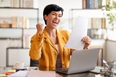 Avrupalı iş kadını elinde kağıtlarla dizüstü bilgisayarını yumrukluyor yazışmaları okuyor, girişimcilik başarısını yurtiçinde kutluyor. Dijital kariyer başarısı, büyük iş haberleri