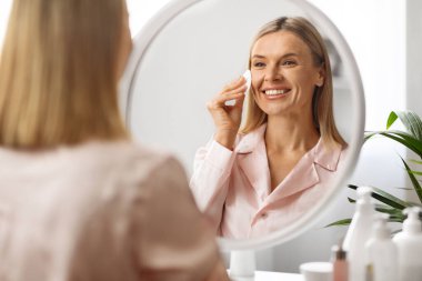 Yüz derisi. Orta yaşlı kadın evde pamuk pedi kullanıyor, gülümseyen olgun bir kadın cildine bakıyor, aynaya bakıyor, evcil güzellik rutininin tadını çıkarıyor, yansımaya odaklanıyor.
