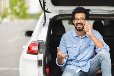 Bin yıllık olumlu Hintli sürücü araba sürerken mola veriyor, bagajda oturuyor, telefonla konuşuyor, el kol hareketi yapıyor ve gülümsüyor.