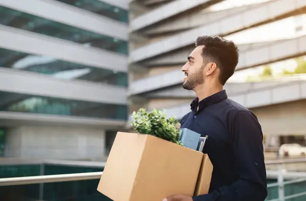 Arab entrepreneur man with belongings box leaving contemporary office center with a smile. Employment challenges and opportunities, start of new career path concept. Free space