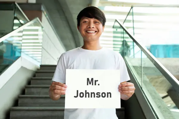 Cheerful friendly millennial asian guy holding placard with name on it, standing on stairs. Chinese guy meeting guest friend or business partner at airport, smiling at camera