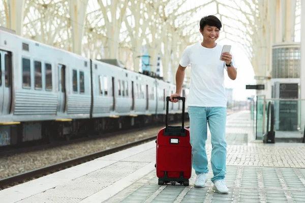 Asian guy traveler using smartphone, walking by train station, checking train schedule online. Young korean man with suitcase travelling by train, using mobile application, buy ticket on Internet