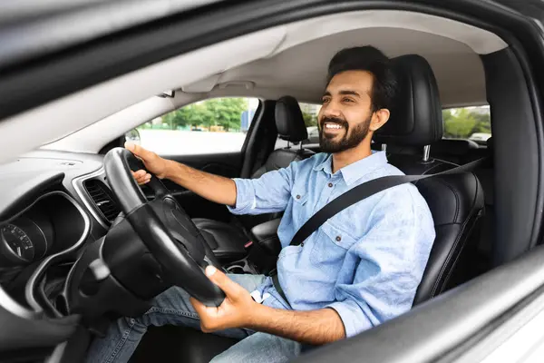 Happy Driver. Side view profile portrait of cheerful positive young indian man sitting in a car on drivers seat. Excited millennial hindu guy riding in the city, holding hands on steering wheel
