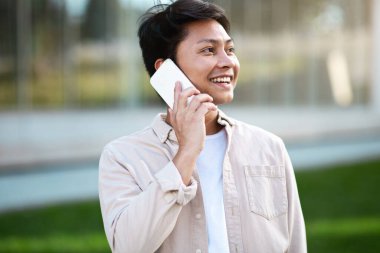 İletişim konsepti. Sıradan kıyafetler giyen yakışıklı Asyalı bir adamın portresi telefon görüşmesi yapıyor, fotokopi odasına bakıyor ve gülümsüyor. Girişimcinin iş görüşmesi var.