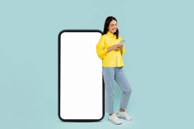 Neşeli, oldukça şık, bin yıllık bir kadın. Mavi stüdyo arka planında beyaz ekranlı büyük bir telefonla poz veriyor. Akıllı telefon, model, reklam için kopyalama alanı, mobil uygulama, online teklif kullanıyor.