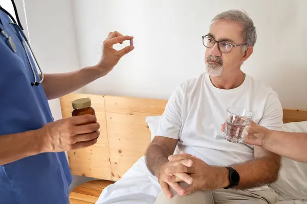看護師は寝室のインテリアで深刻なシニアの白人男性に錠剤を与えます 医薬品 ビタミン 鎮痛剤 ガラス水などの問題に対する医療支援 — ストック写真