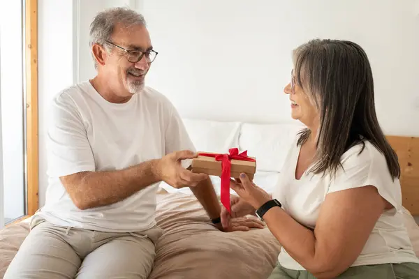 Glad old caucasian lady gives gift box to man, sit on bad in bedroom interior. Celebration birthday, love, anniversary surprise, holiday together and congratulate at home