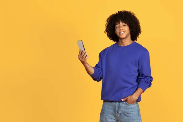 快乐的黑人小伙子用手机笑着对着相机笑 快乐的千年黑人男人手里拿着智能手机 站在黄色工作室的背景上 复制空间 — 图库照片